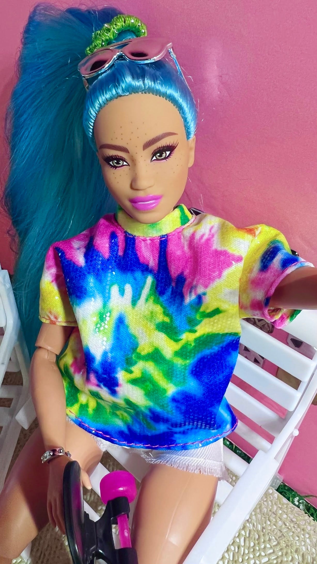 Doll with blue hair and tie-dye shirt sitting on a chair against a pink background