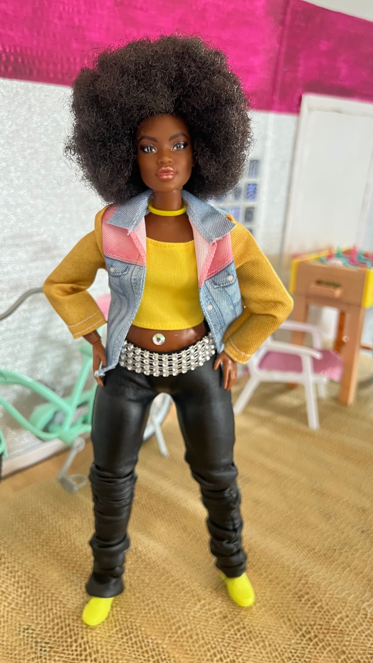 Doll with an afro hairstyle wearing a yellow top, denim jacket, and black pants in a room setting.